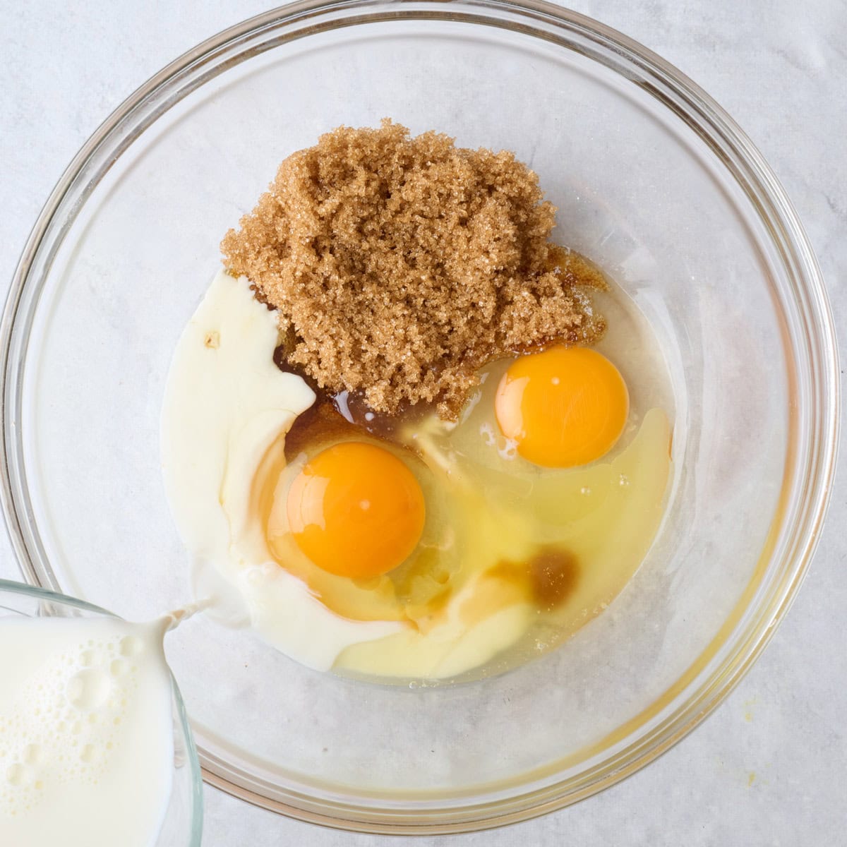 Eggs, brown sugar, and vanilla in a bowl with milk being poured in.