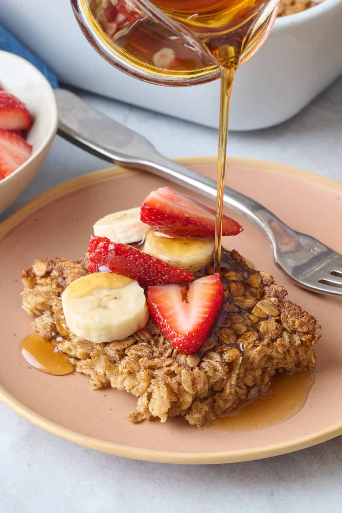 Baked oatmeal on a plate topped with sliced bananas and strawberries with maple syrup being poured on top.