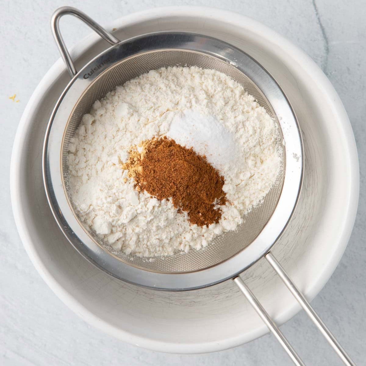 Dry ingredients in a sifter over a bowl before being sifted.
