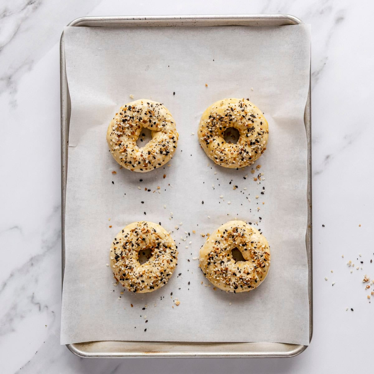 4 bagels on small parchment lined baking sheet with egg wash and everything bagel seasoning before baking.