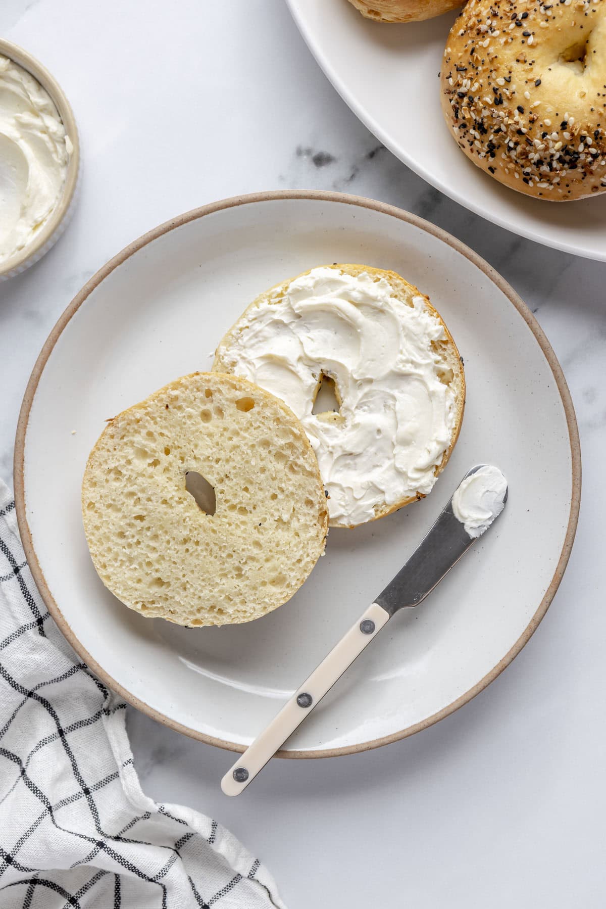 Cottage cheese bagel on a plate, split in half with cream cheese spread on one half, plate of more bagels nearby.