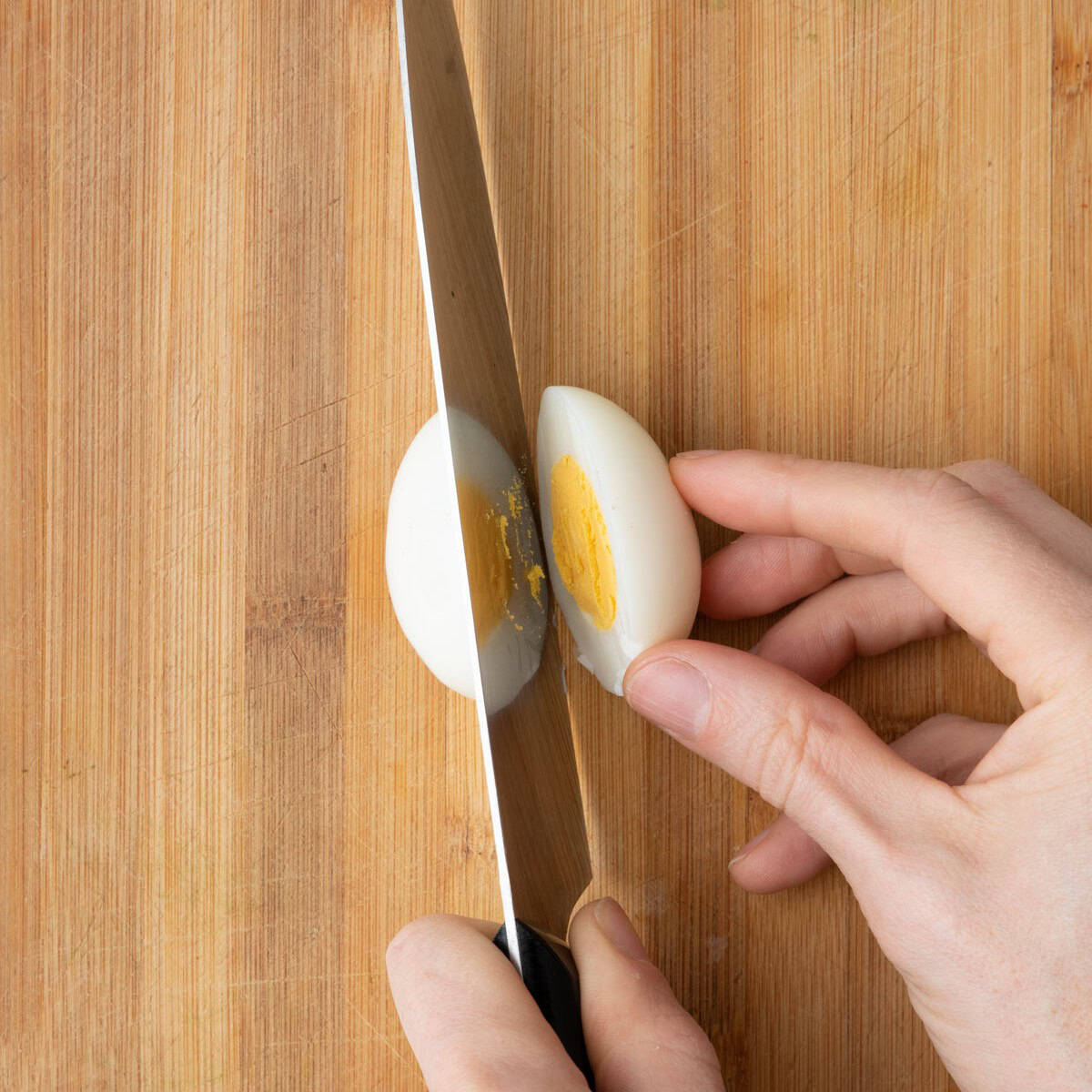 Knife slicing a boiled egg in half lengthwise on a wooden cutting board.