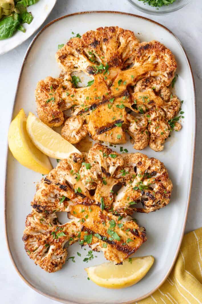 Cauliflower steaks on a small platter with a small dish of chopped parsley, garnished with fresh parsley and lemon wedges.