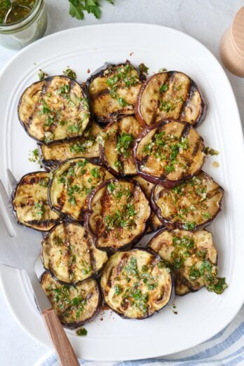 Grilled eggplant slices on a platter with sauce spooned on top, serving set and jar or small bowl of extra sauce with spoon dipped inside nearby.