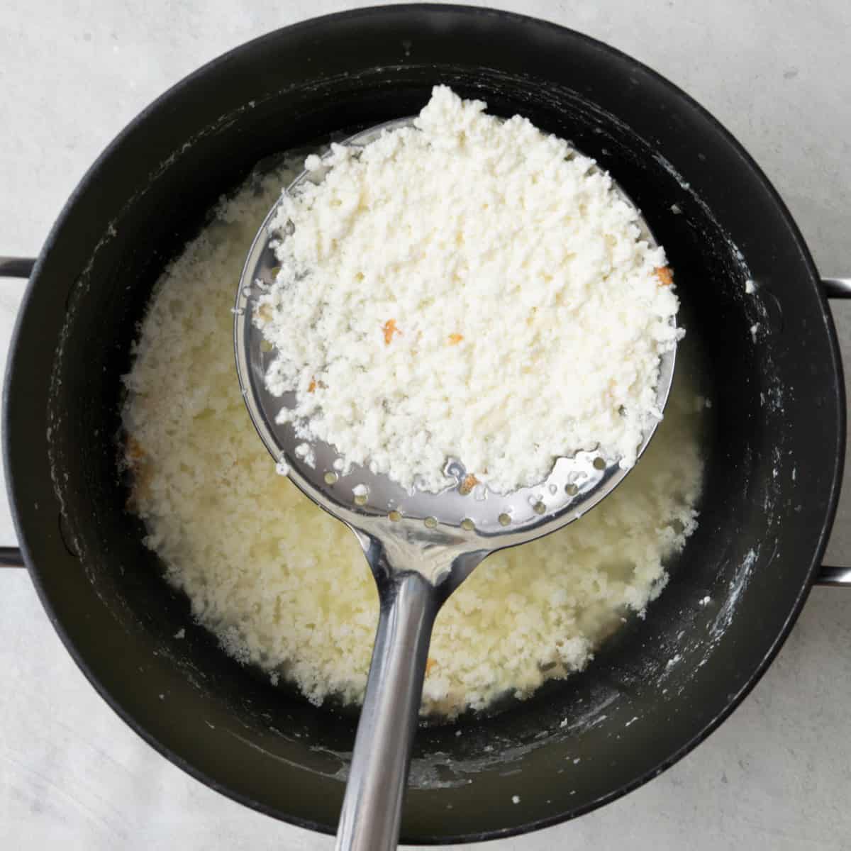 Slotted spoon removing clotted cream from the surface of the saucepan, showing liquid below it.