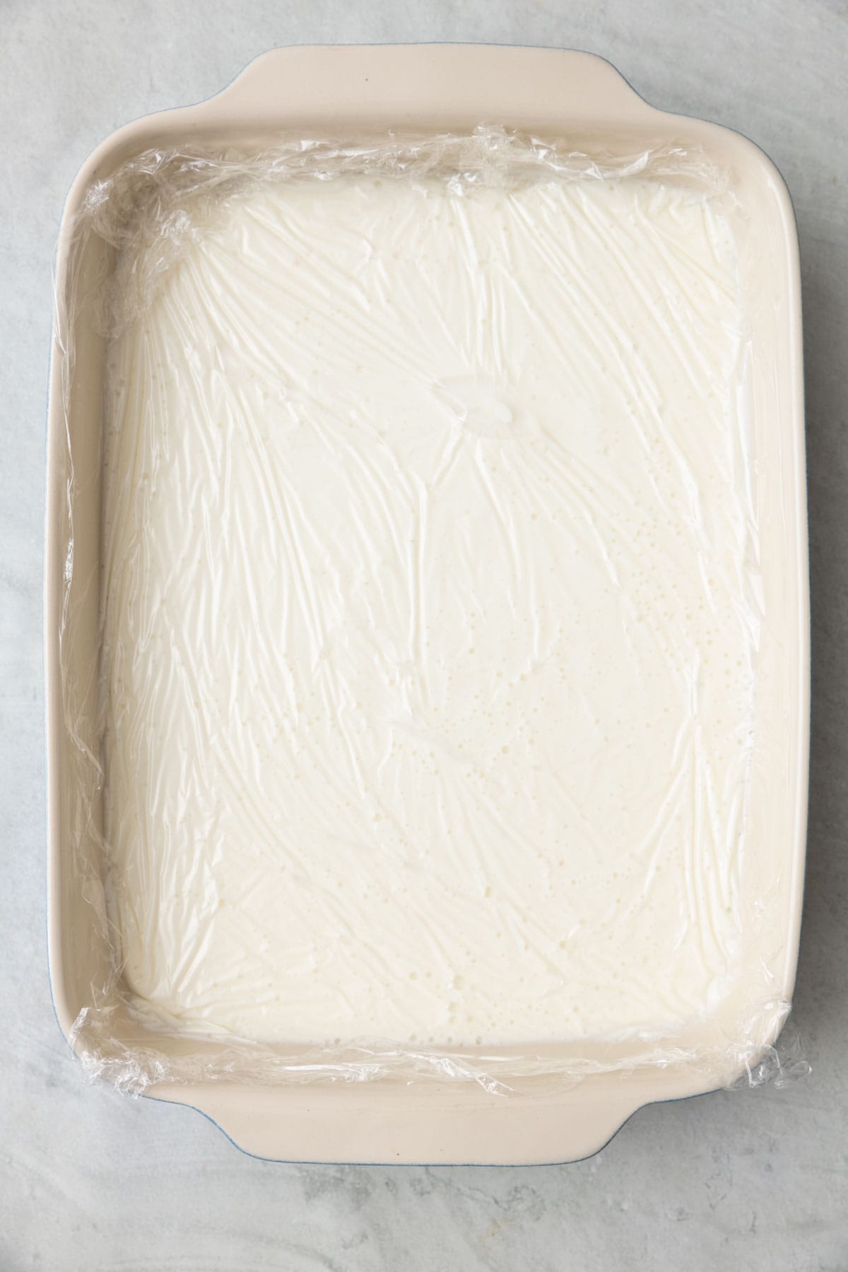 Ashta in casserole dish with a piece of plastic wrap pressed directly on the surface.