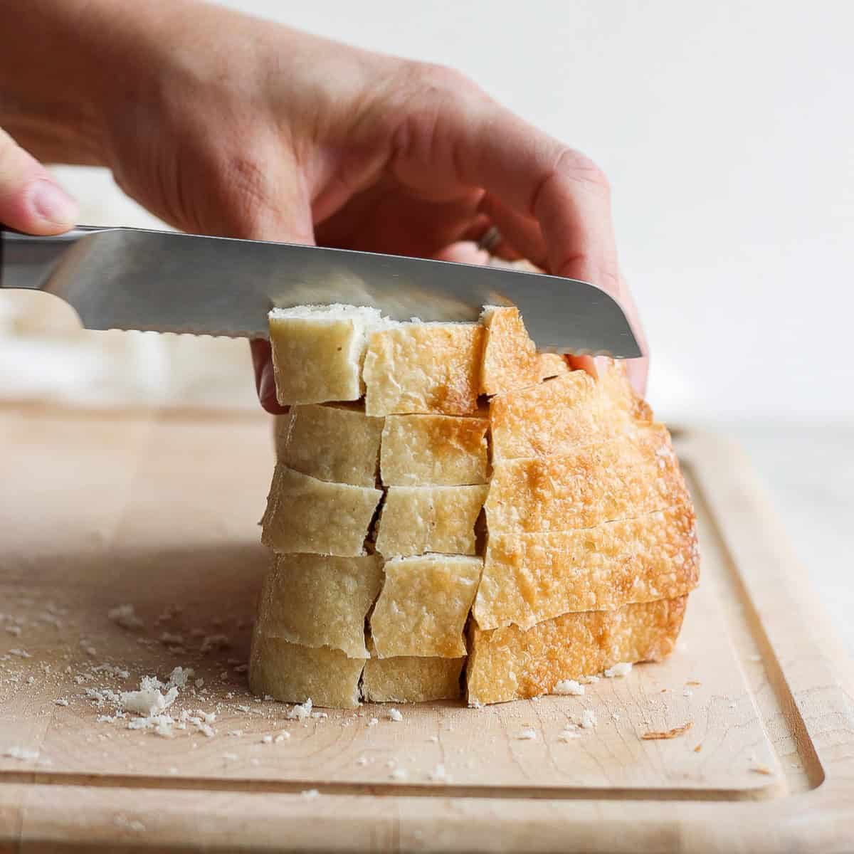 Stack of bread slices on a cutting board cut into strips and being cut into cubes.