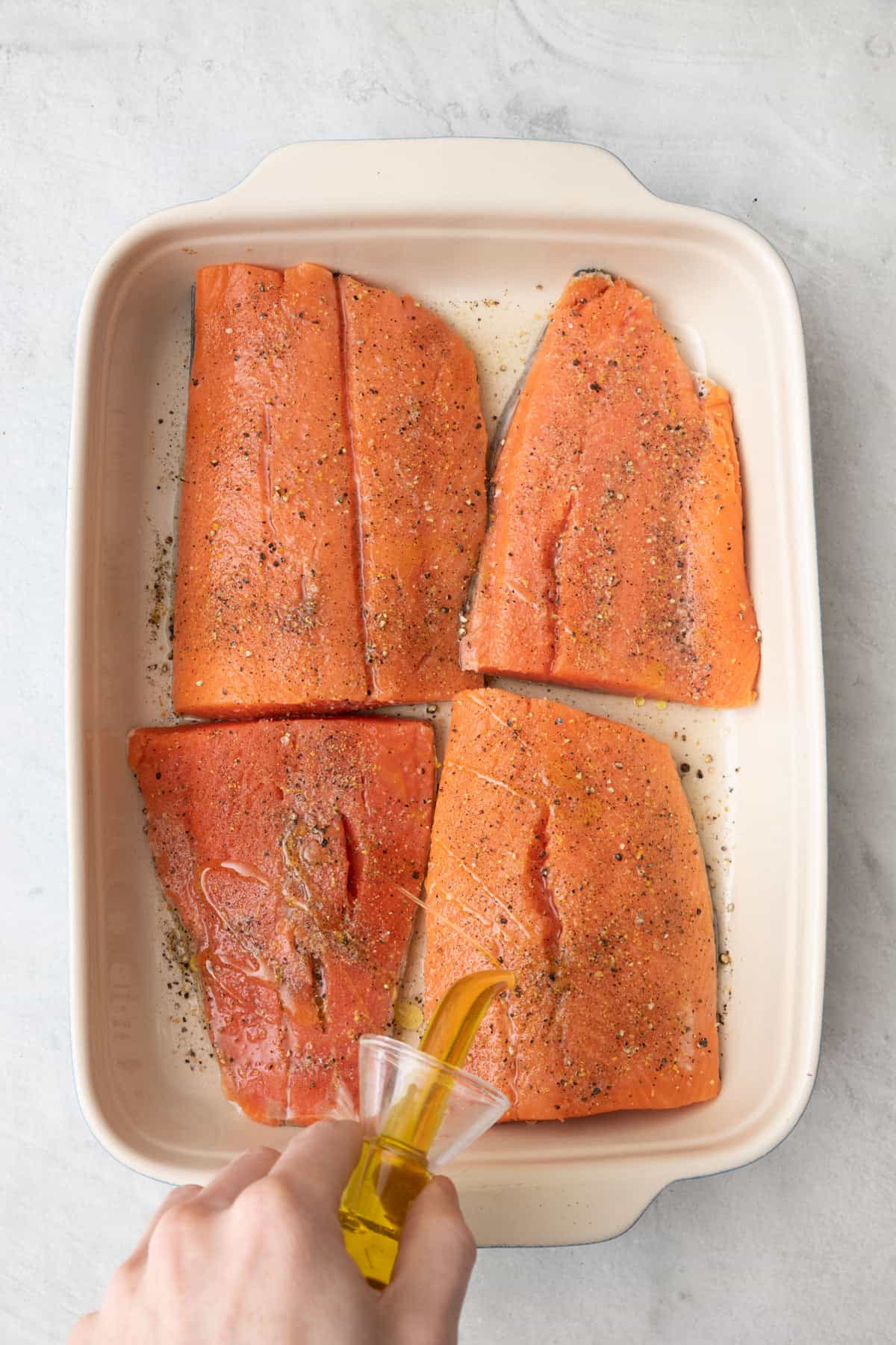 Oil being poured over 4 salmon fillets in an 8 x 11 in baking dish with seasoning and lemon juice added.