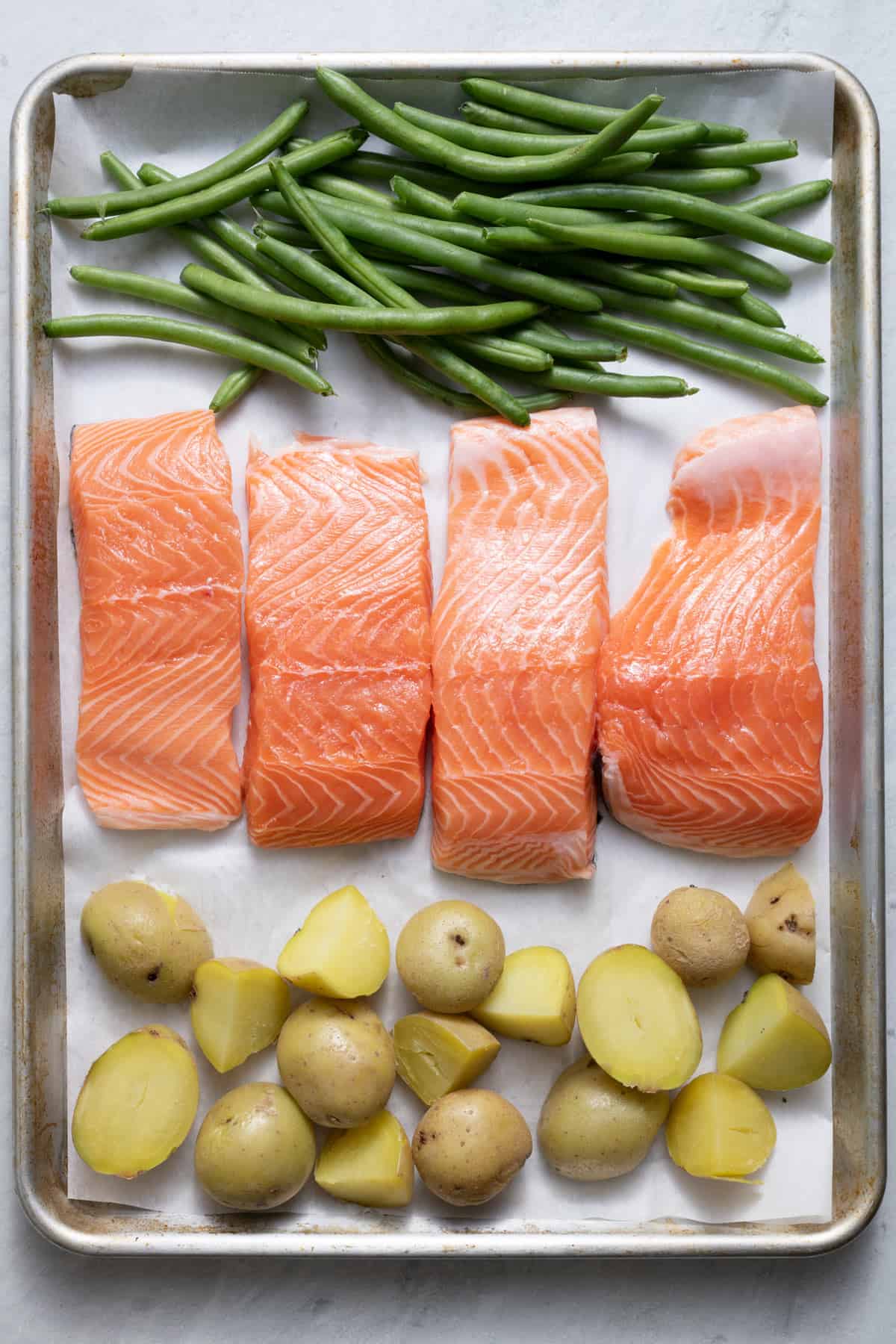 Halved cooked new potatoes, four salmon fillets, and fresh green beans on a parchment lined sheet pan.
