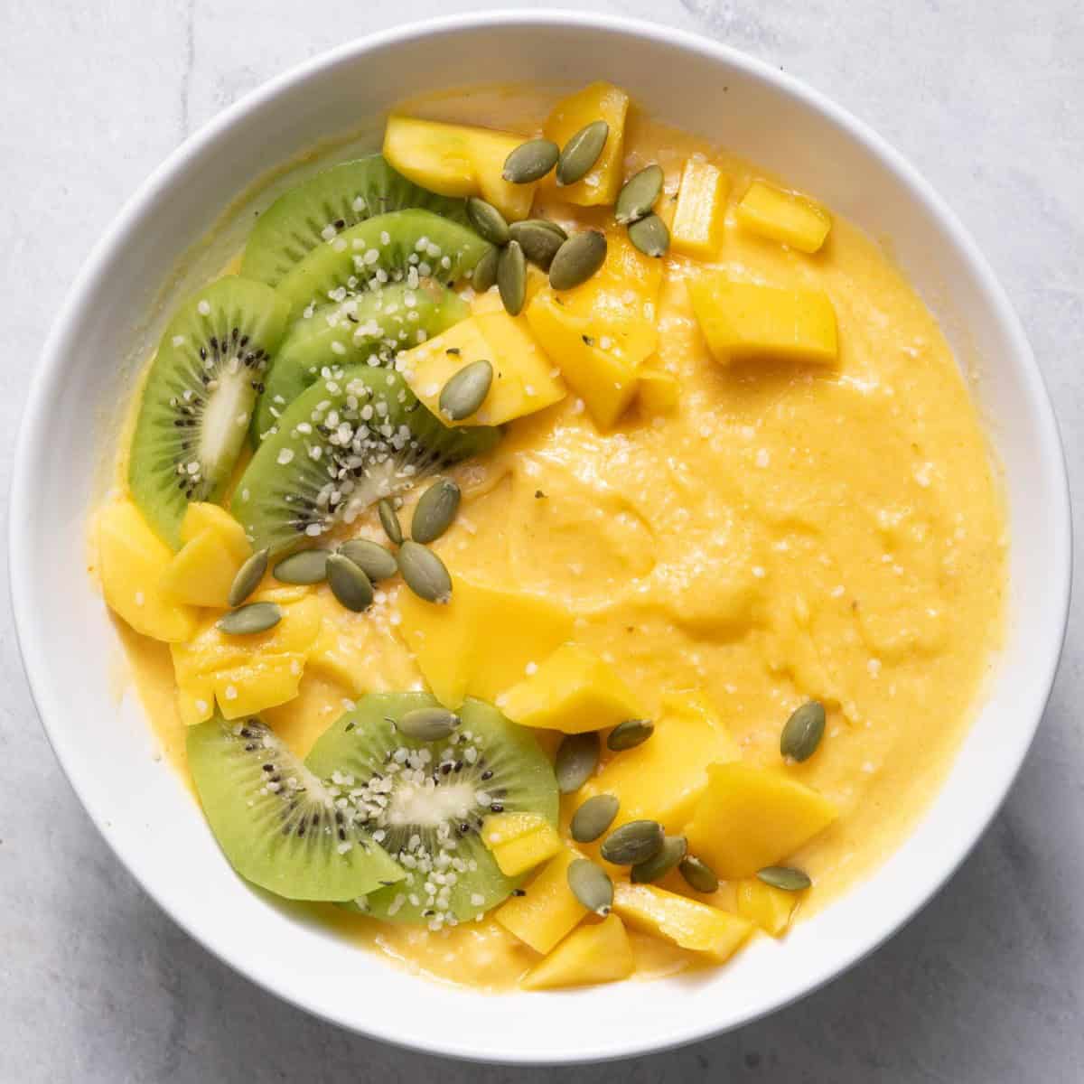 Mango banana smoothie bowl topped with sliced kiwi, fresh mango, and sunflower seeds..