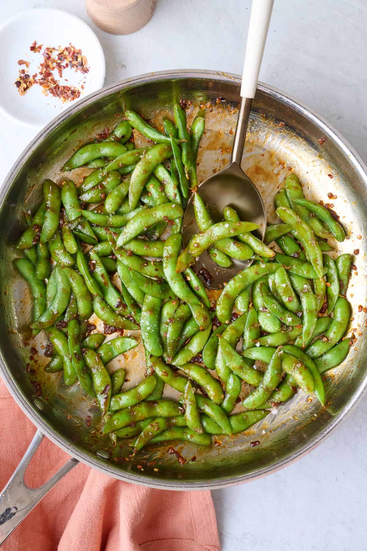 Spicy edamame in skillet garnished with flaky sea salt, serving fork or spoon resting in pan, small dish of red pepper flakes nearby.