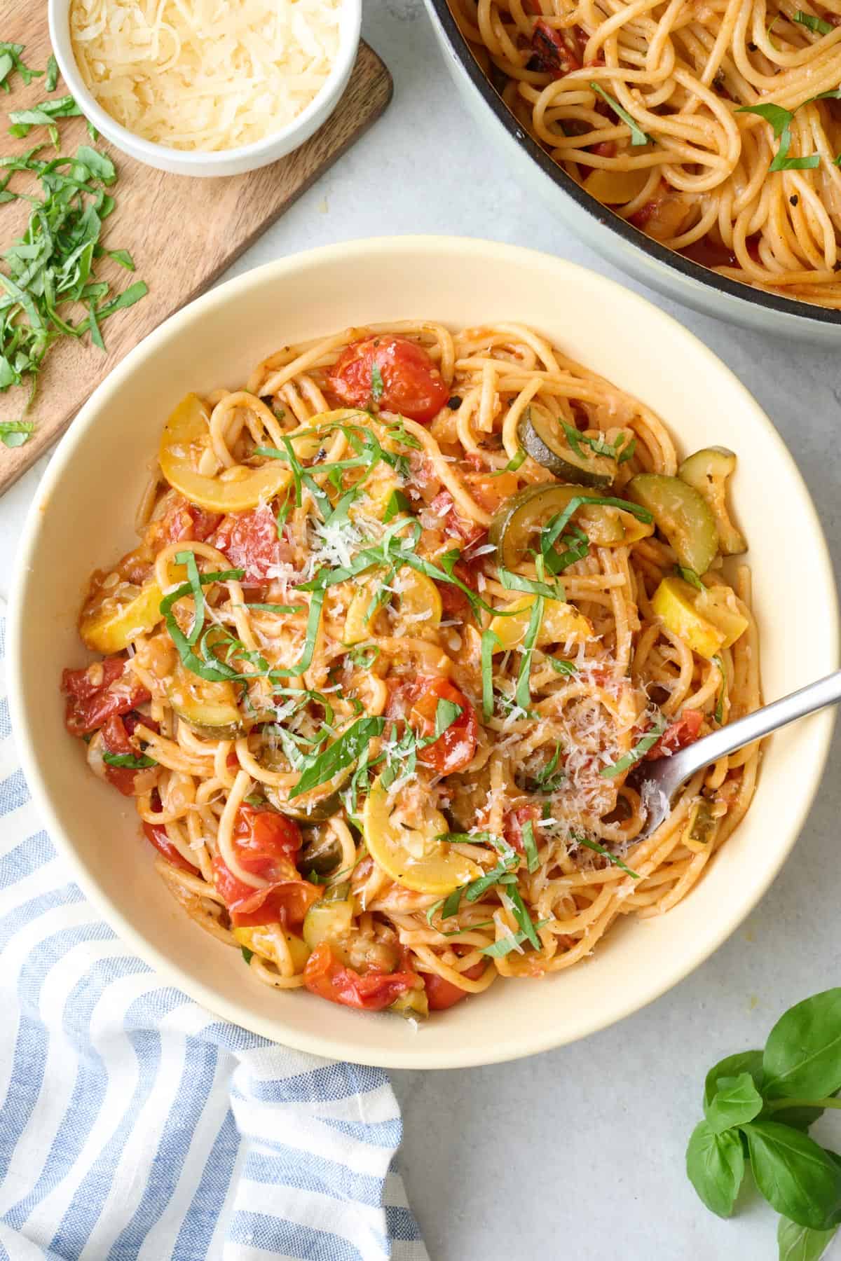 Serving of veggie spaghetti in a shallow pasta bowl with pot and parmesan bowl nearby.