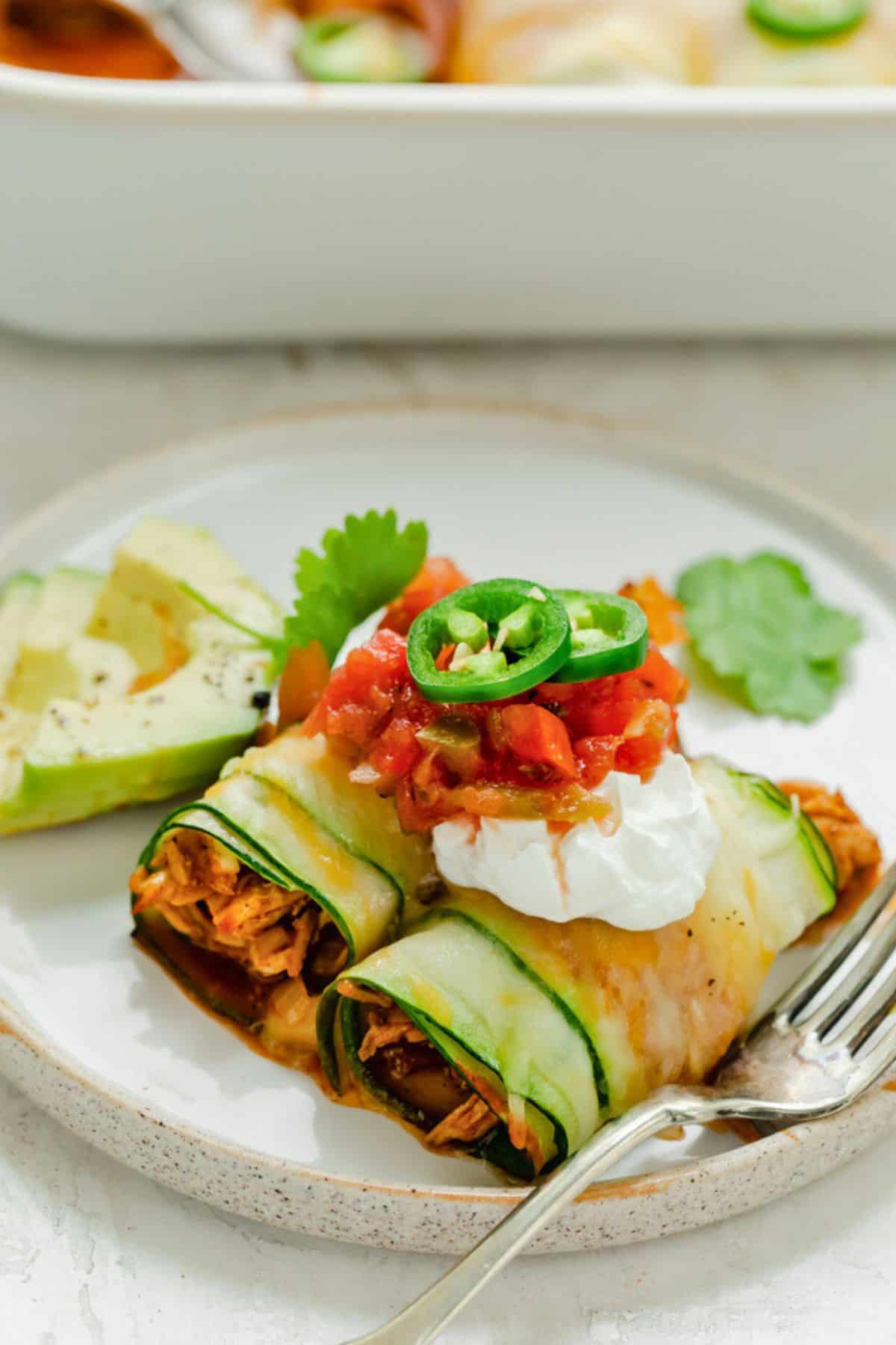 Two pieces of zucchini enchilada on a plate topped with salsa and jalapenos.