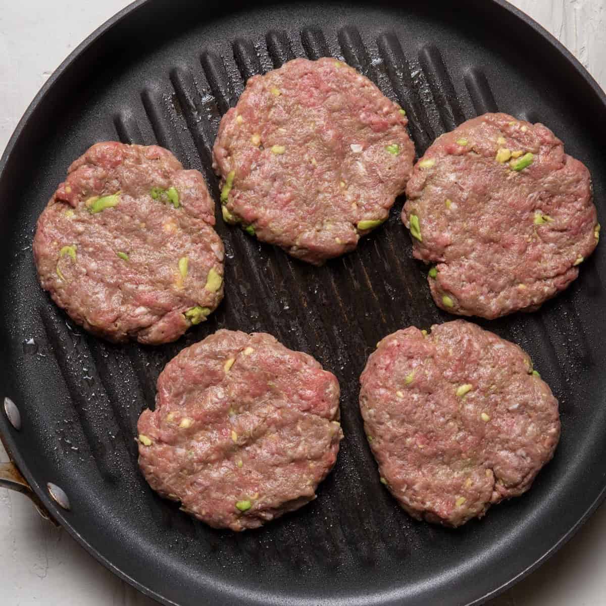 Burger patties with avocado chunks on a grill pan.