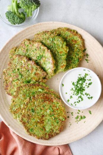 Broccoli fritters on a round plate garnished with a small dish of greek yogurt sauce with chopped parsley on top.