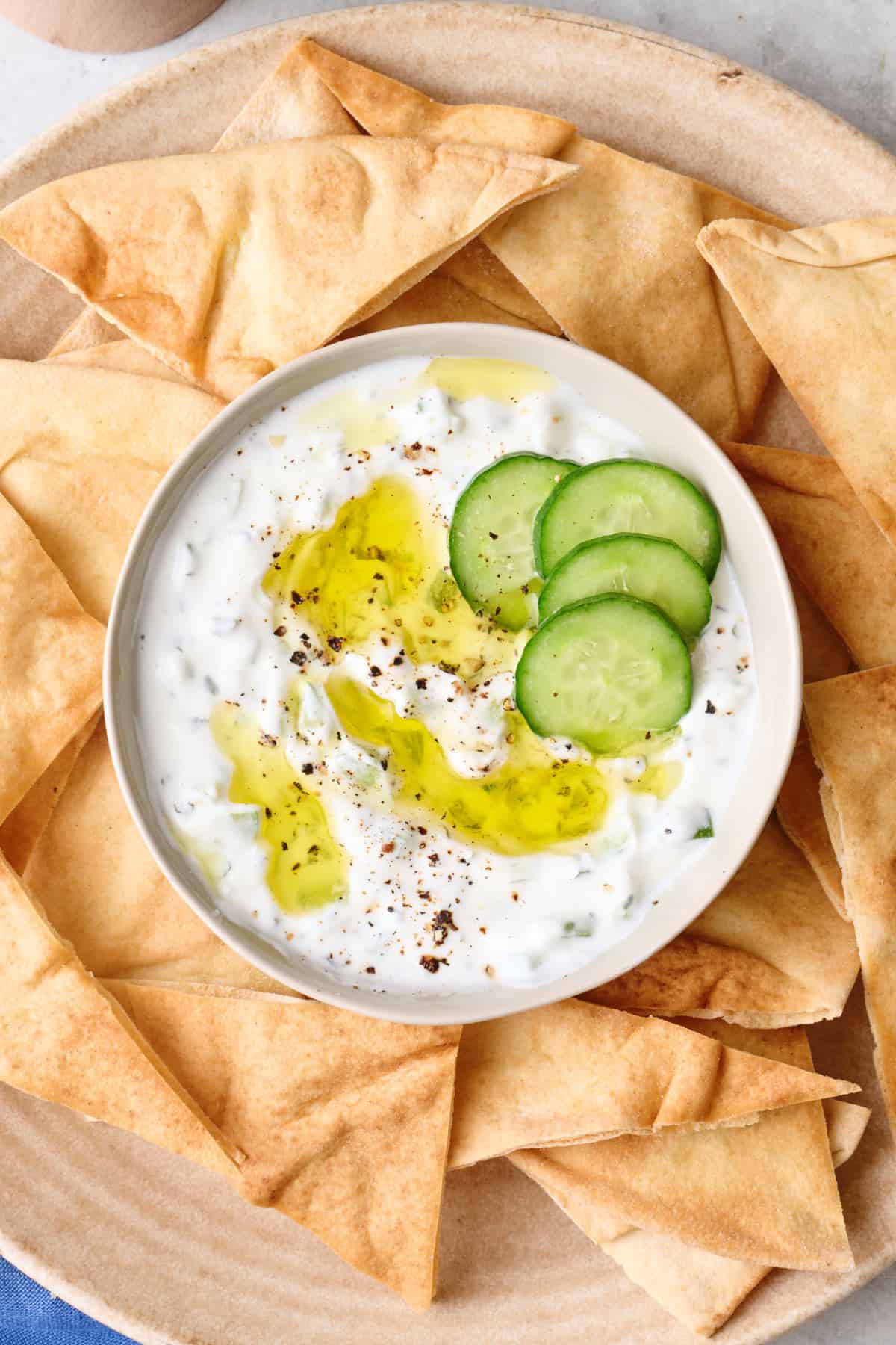 Cucumber yogurt sauce topped with oil and sliced cucumbers on a plater with pita chips.