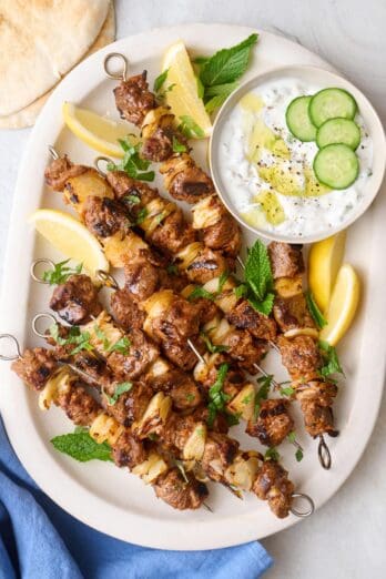 Grilled Lamb kabobs on a platter with a side of yogurt sauce and lemon wedges and lots of fresh mint.