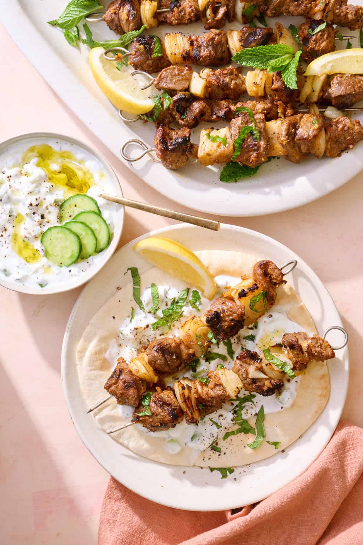 Grilled lamb kabobs recipe with two kabobs on a pita with yogurt sauce and platter of more skewers nearby.