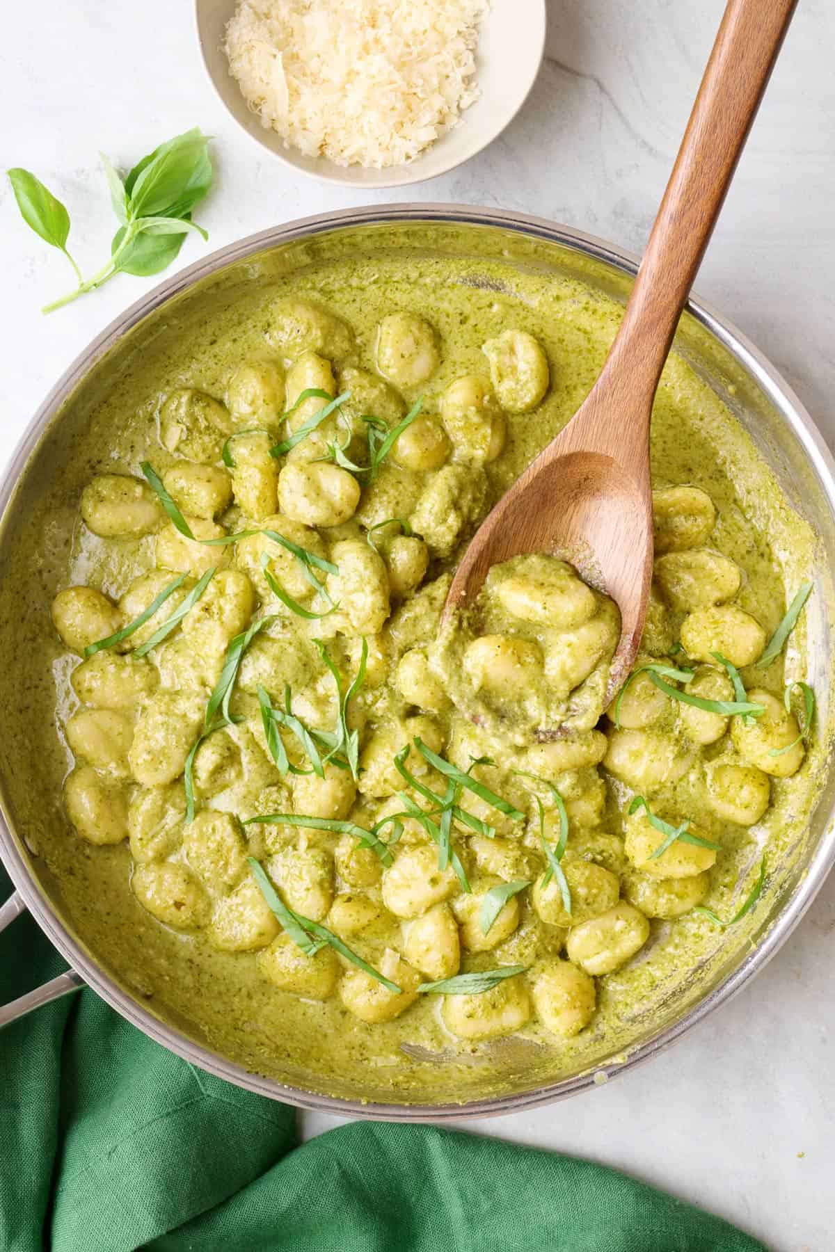 Creamy pesto gnocchi in skillet with serving spoon dipped inside pan, small dish of parmesan nearby.