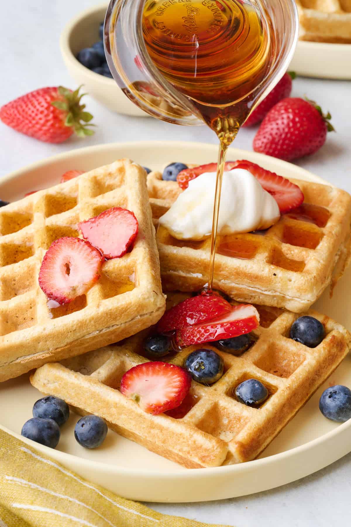 Plate of homemade protein waffles topped with fresh fruit with maple syrup being poured on top.