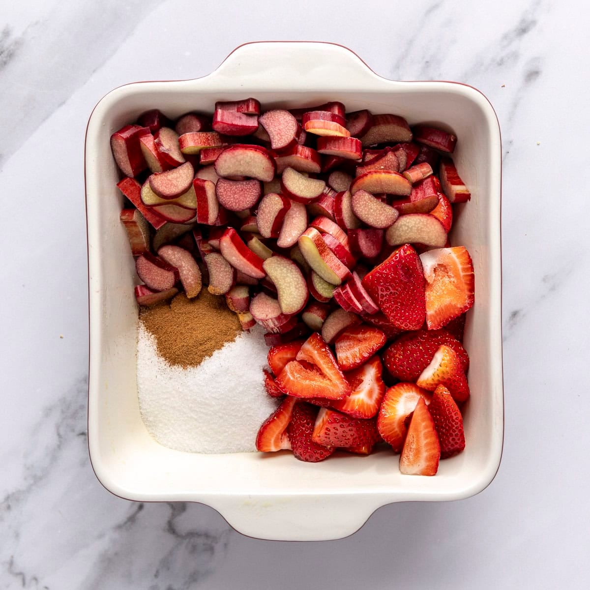 Chopped rhubarb, strawberries, sugar, and cinnamon in a square baking dish before tossing together.