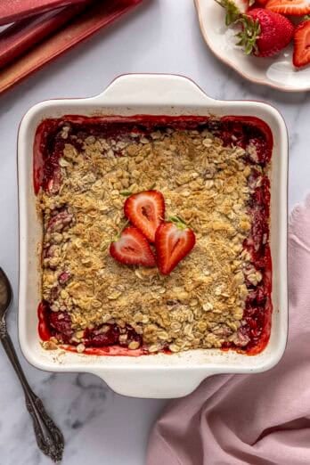 Strawberry rhubarb crisp in baking dish a few slices of fresh strawberries on top and with extra fruit and serving spoon nearby.