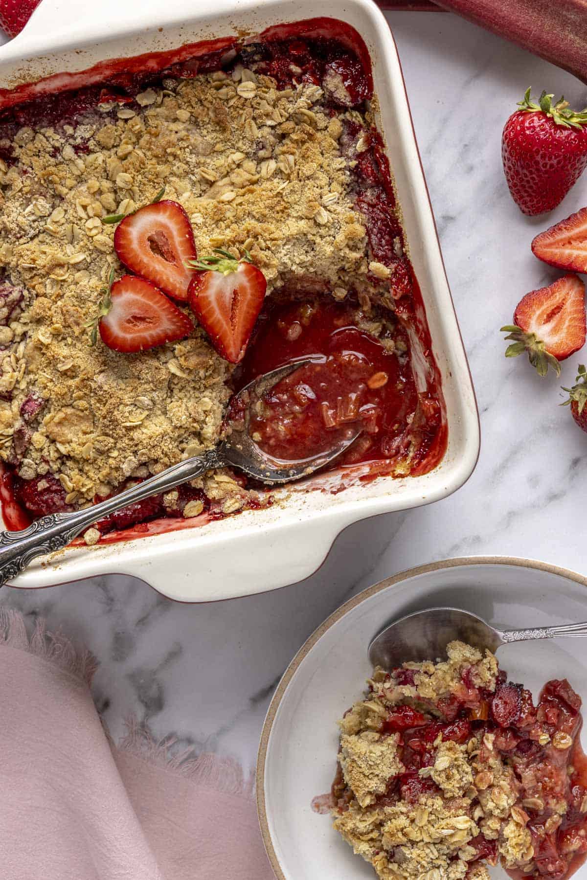 Serving of warm strawberry rhubarb crisp with serving removed to a nearby plate and serving spoon dipped inside baking dish.