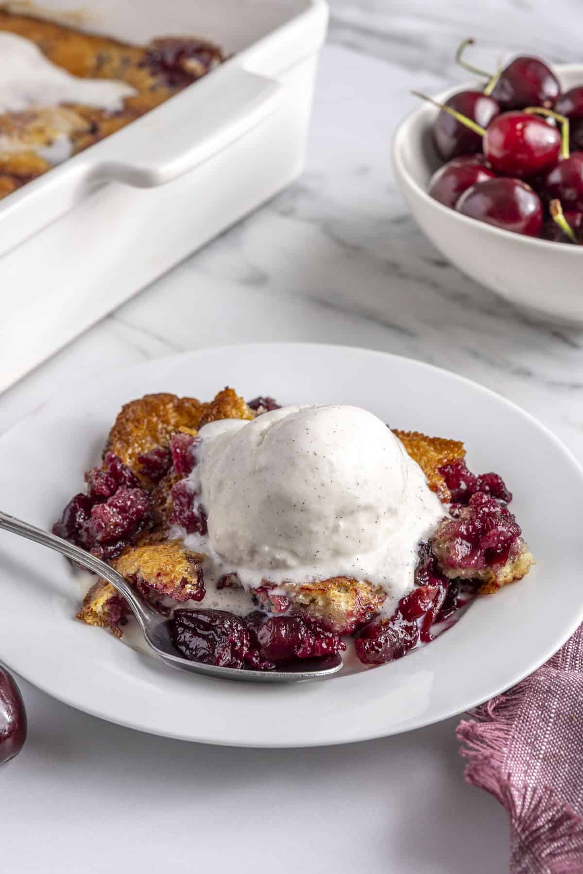 Serving of cherry cobbler served with a scoop of ice cream, spoon on plate, baking dish and bowl of cherries nearby.