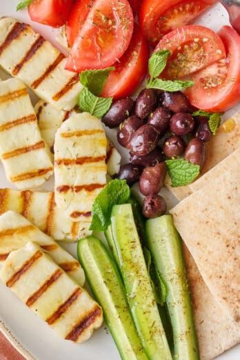 Grilled halloumi recipe on platter with fresh veggies, olives, and pita.
