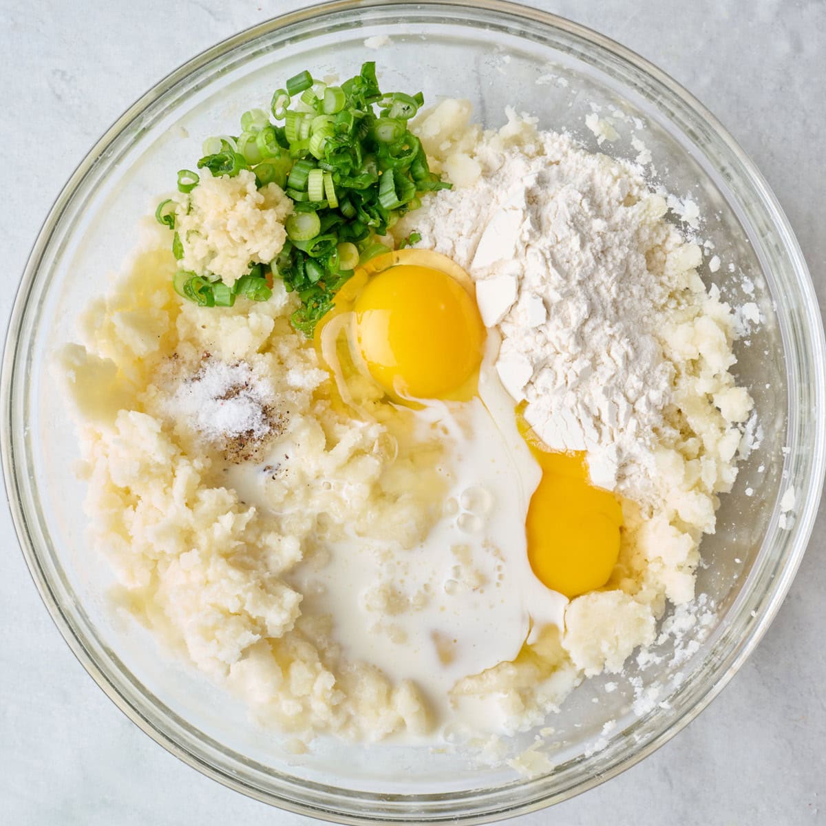 Mashed potatoes, eggs, milk, seasoning, garlic, green onions, and flour in a bowl before mixing.