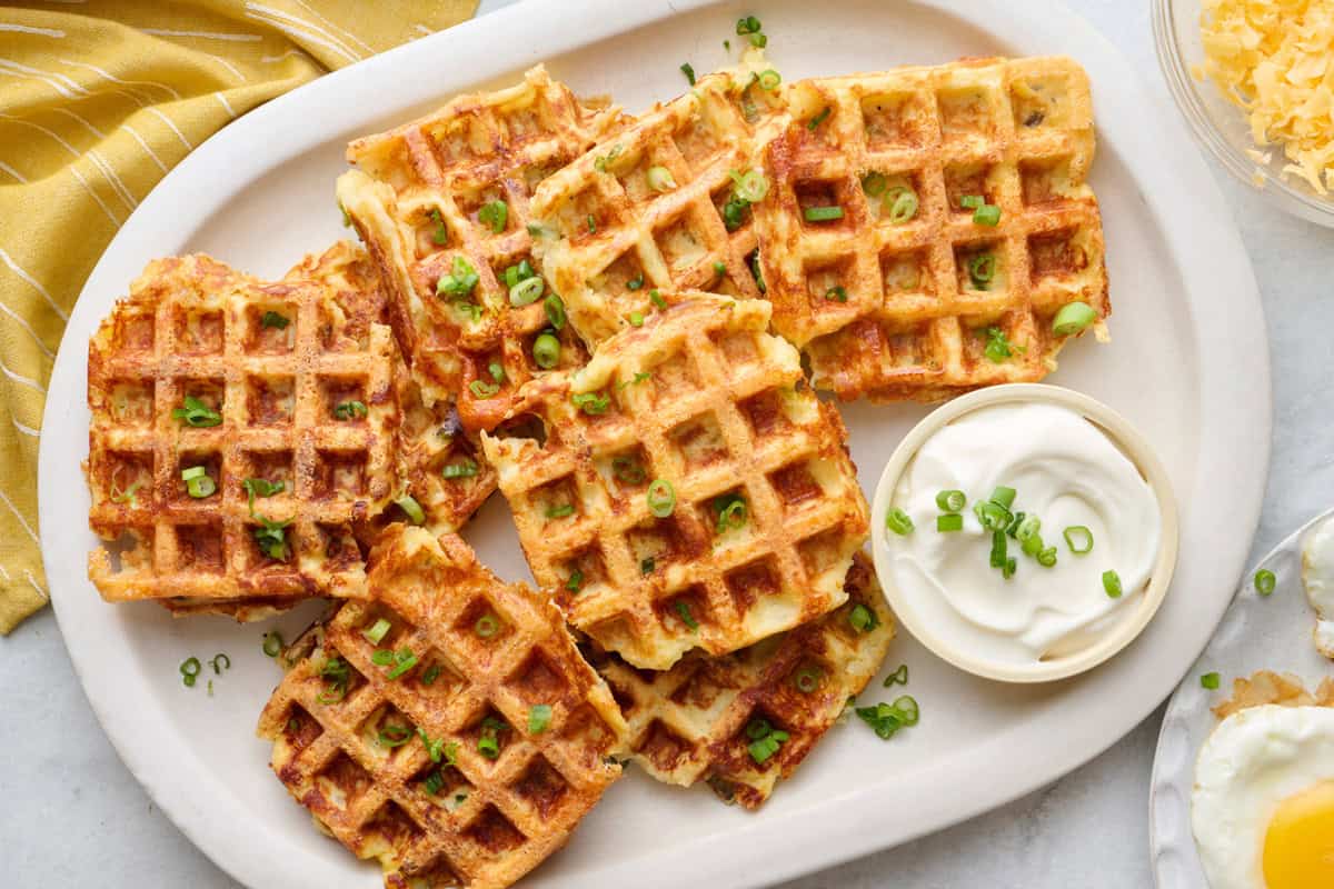 Crispy potato waffles on a platter garnished with chopped green onions, with a small dish of sour cream and small plates of cooked eggs nearby.