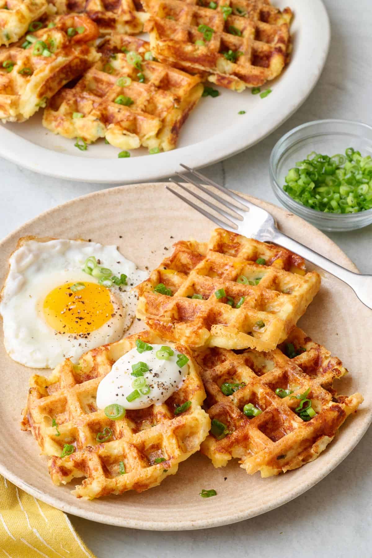 Plate of cheesy mashed potato waffles on a plate topped with sour cream and green onions, served with a sunny side up egg.