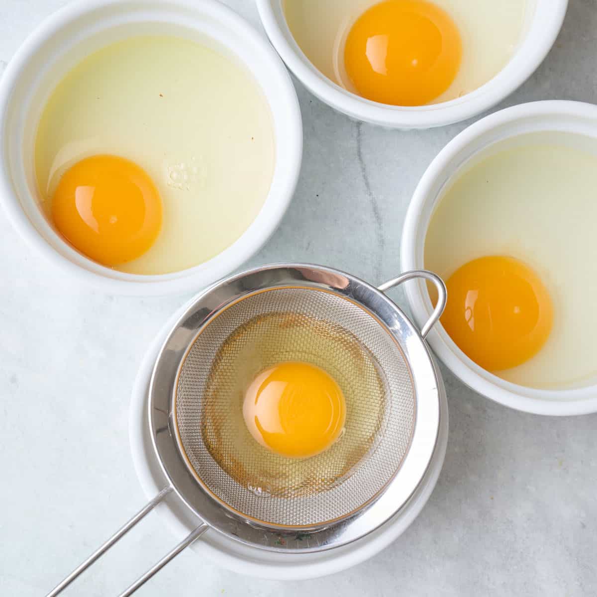 One cracked egg in mesh sieve over a small bowl with 3 others already drained in small bowls.