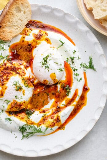 Two bowls of Turkish eggs served over yogurt, topped with hot chili butter and garnished with fresh dill, crust bread nearby