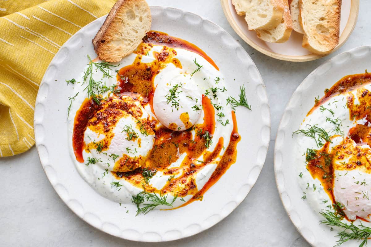 Two bowls of Turkish eggs served over yogurt, topped with hot chili butter and garnished with fresh dill, crust bread nearby