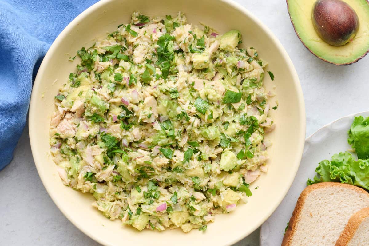 Avocado tuna salad in a serving bowl with a few slices of bread, leafy lettuce and half avocado nearby.