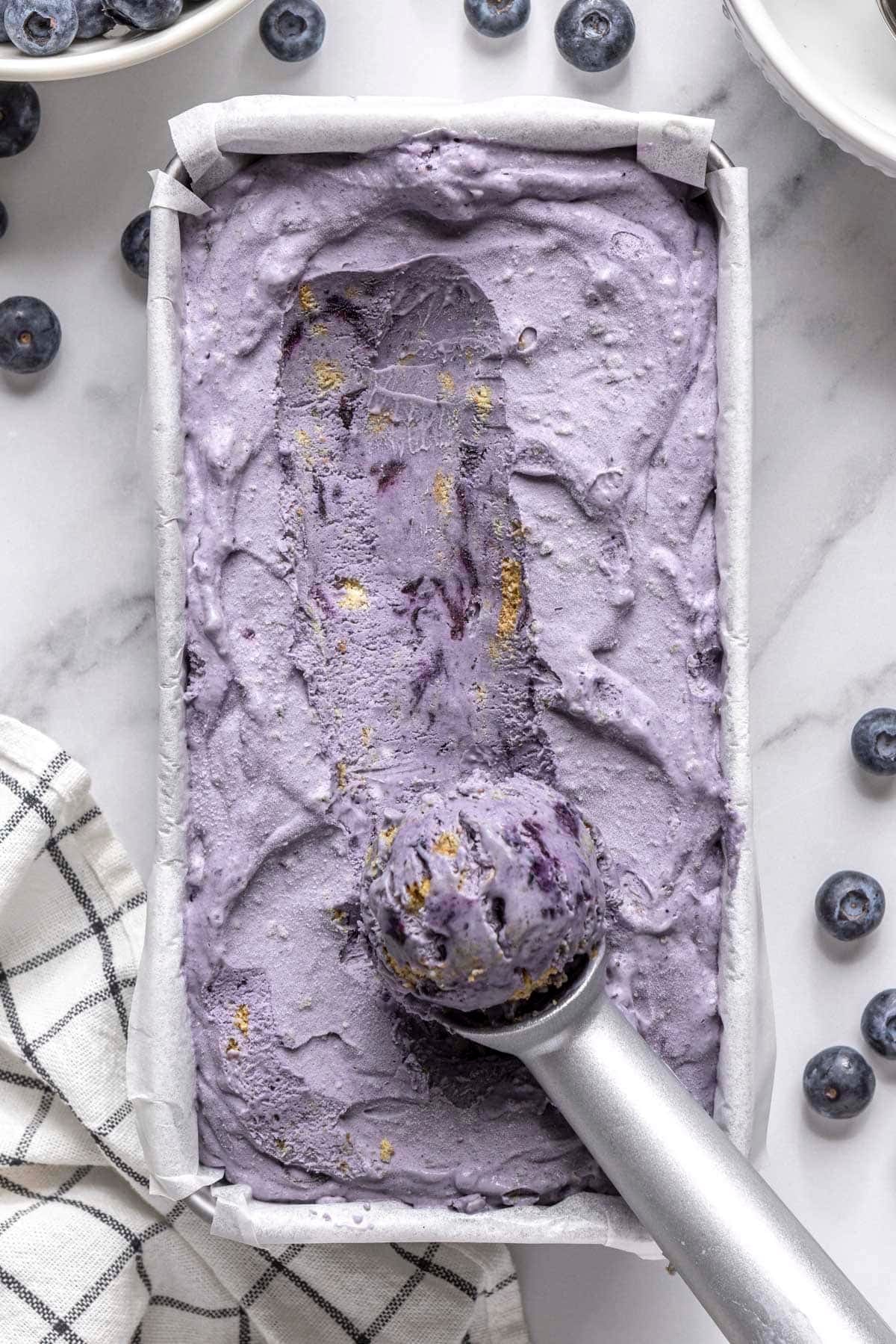 Blueberry ice cream with graham crackers in a dish with ice cream scoop resting on top with some ice cream on it.