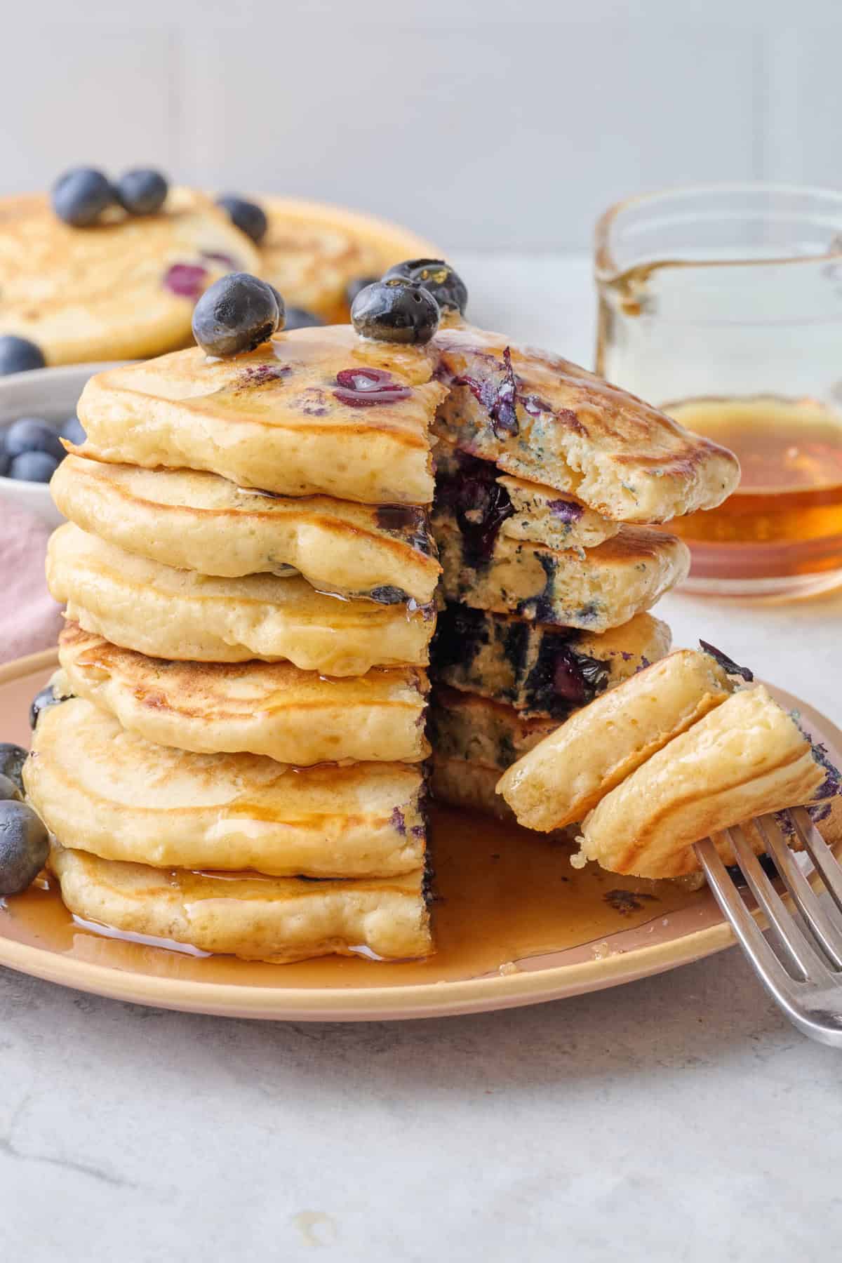 Stack of homemade blueberry pancakes covered in syrup with a bite cut out and fork with pancake resting on plate.
