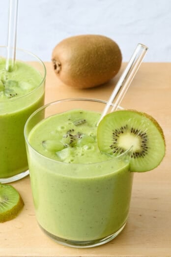 2 glasses of kiwi smoothie with a few kiwi chunks on top and slice kiwi on rim, clear reusable straws dipped inside.