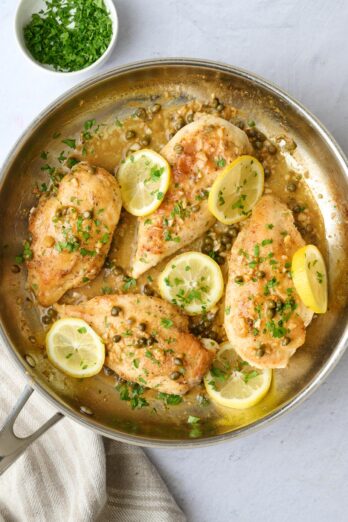 Lemon chicken piccata in skillet with capers and sauce spooned over chicken, garnished with chopped parsley and fresh sliced lemons on top.