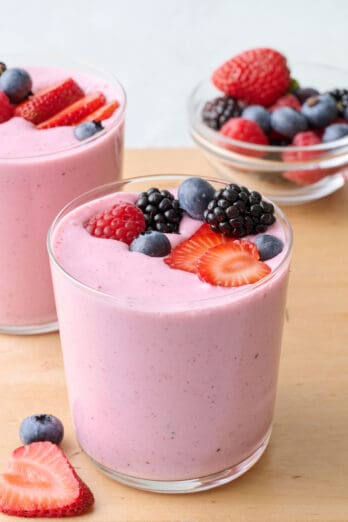 2 glasses of mixed berry smoothie topped with extra mixed berries and more berries either in a dish nearby.