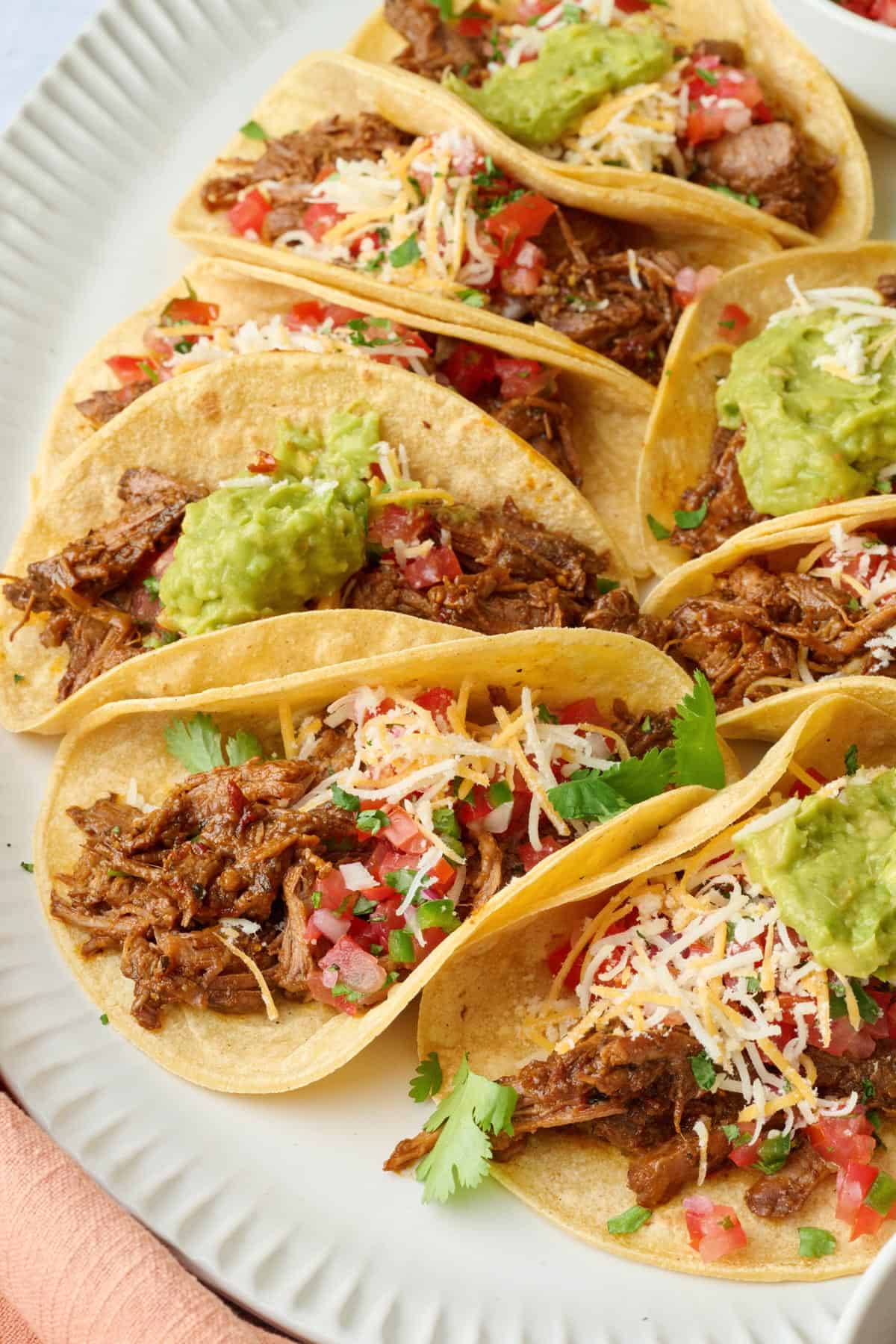 Shredded beef tacos on a platter topped with fresh pico de gallo, guacamole, shredded cheese.