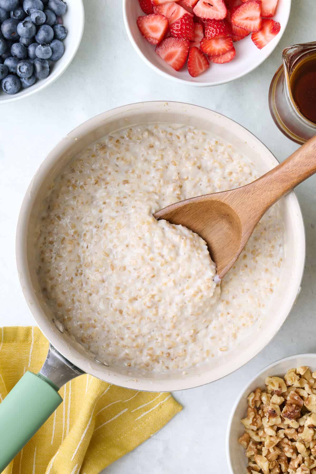 Steel cut oatmeal in pot with wooden spoon dipped inside, toppings in small dishes nearby.