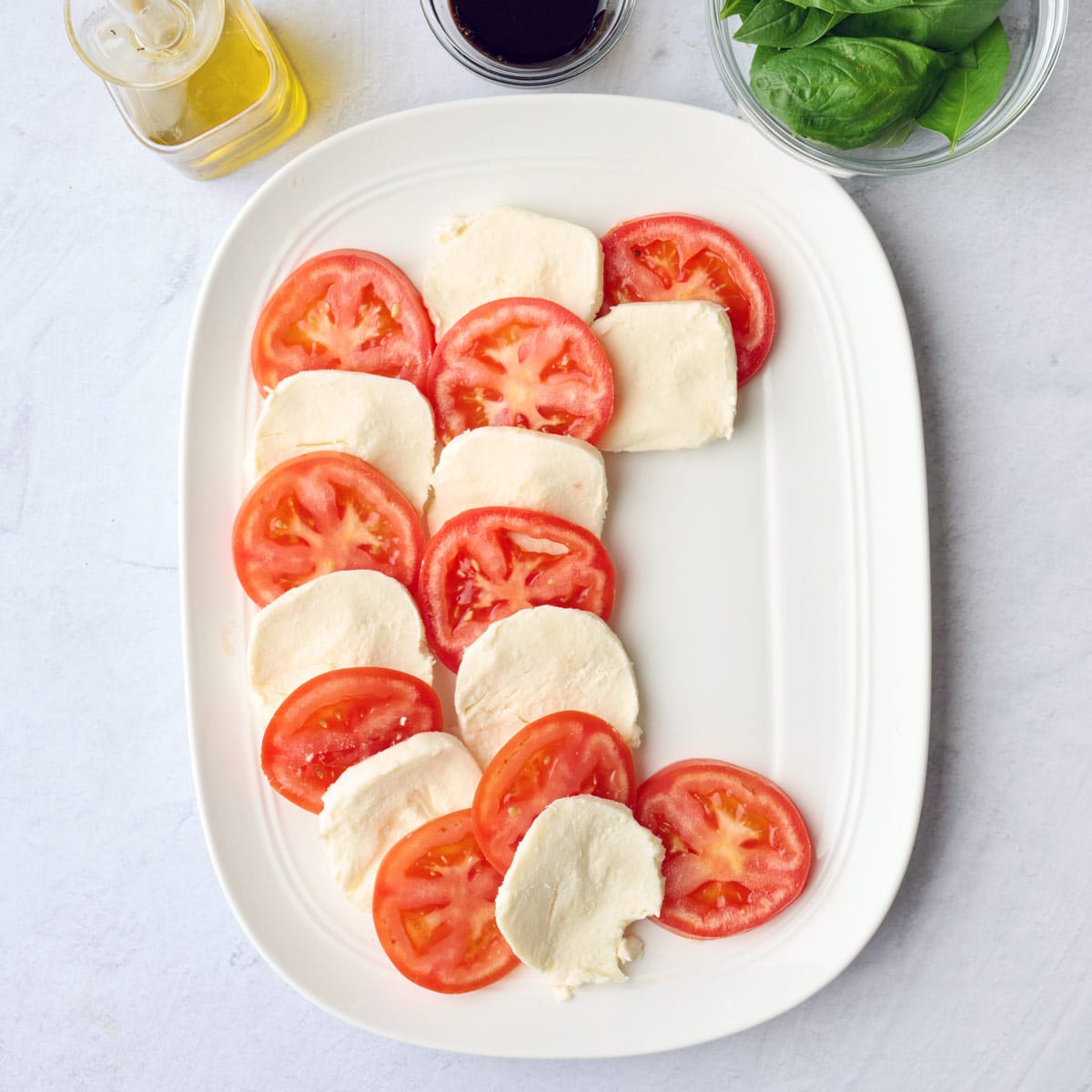 About ยพ of plate assembled with alternating slices of tomato and mozzarella, small dish of basil leaves nearby.
