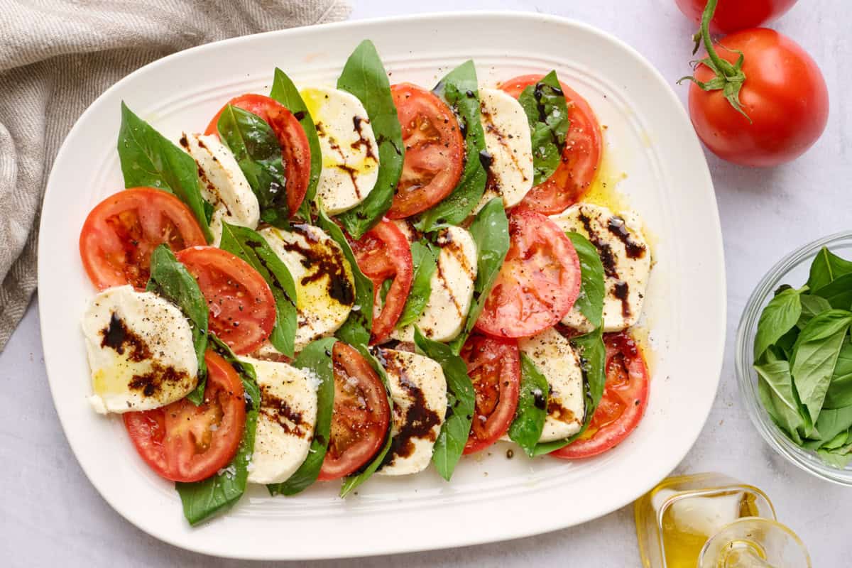 Assembled caprese salad on an oval platter, seasoned with flaky salt and pepper and a drizzle of oil and balsamic glaze on top.