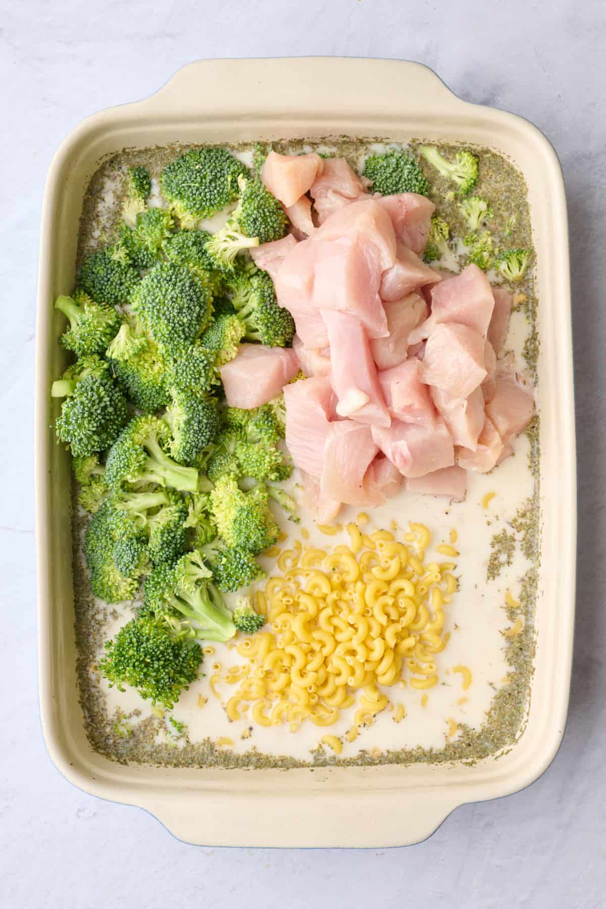 Pasta, chicken cubes, and broccoli florets added to baking dish with sauce ingredients.
