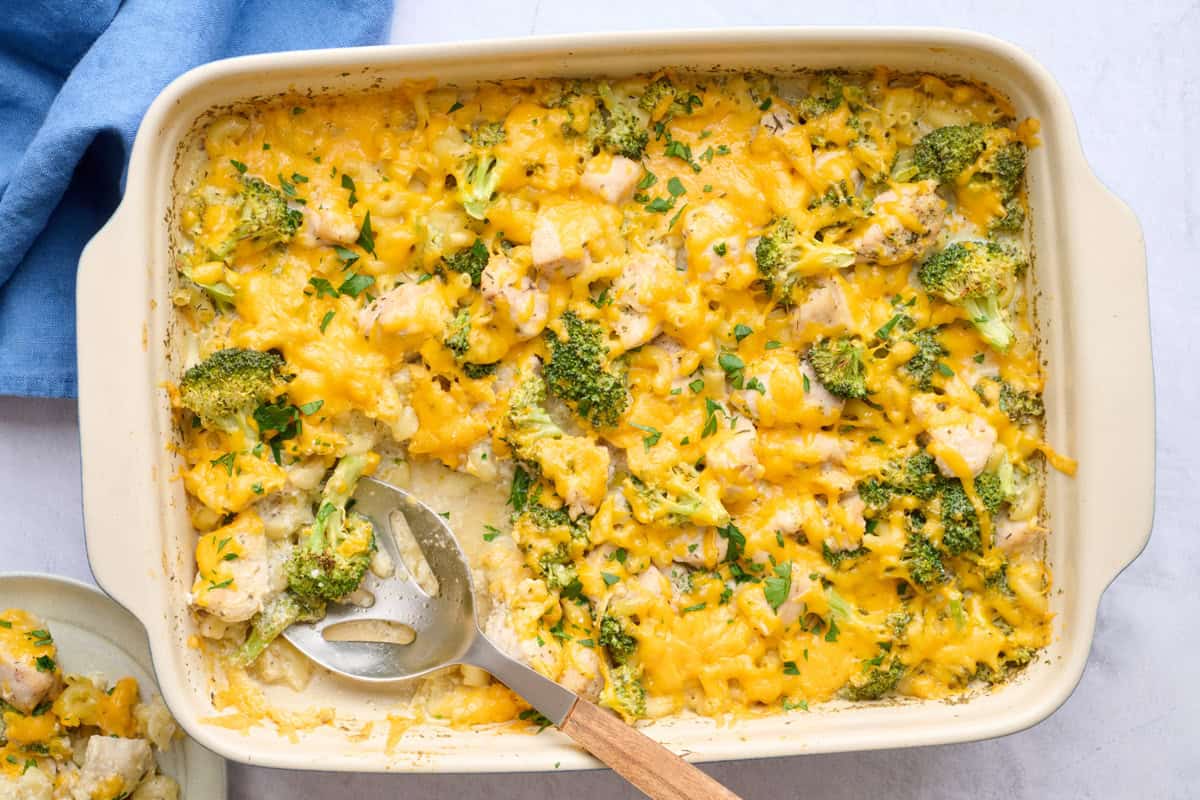 Casserole dish of Chicken broccoli bake garnished with chopped parsley with a serving removed to a small plate nearby by and serving spoon dipped inside dish.