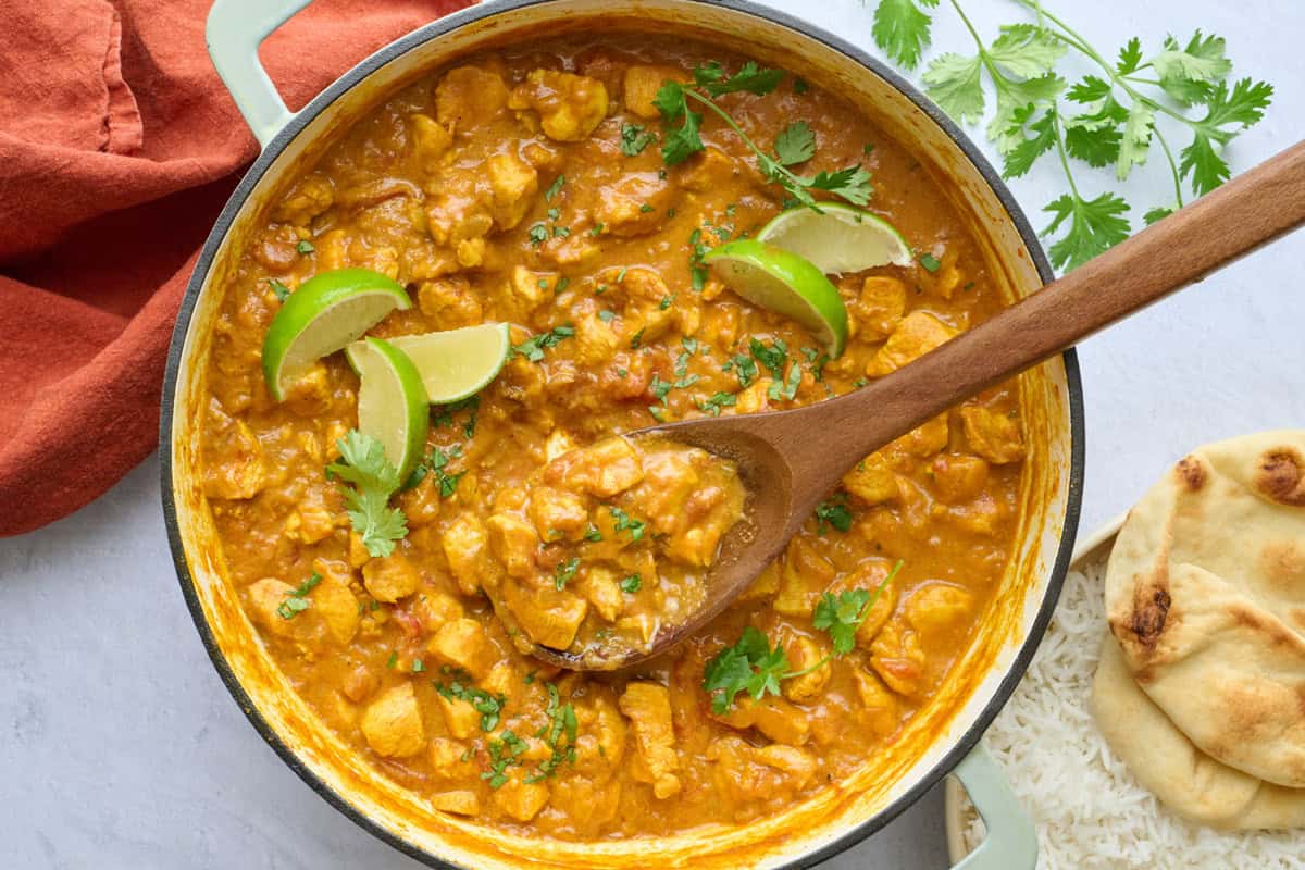 Coconut curry chicken in pot with spoon dipped inside, garnished with cilantro, lime wedges, bowl of rice, and naan nearby.