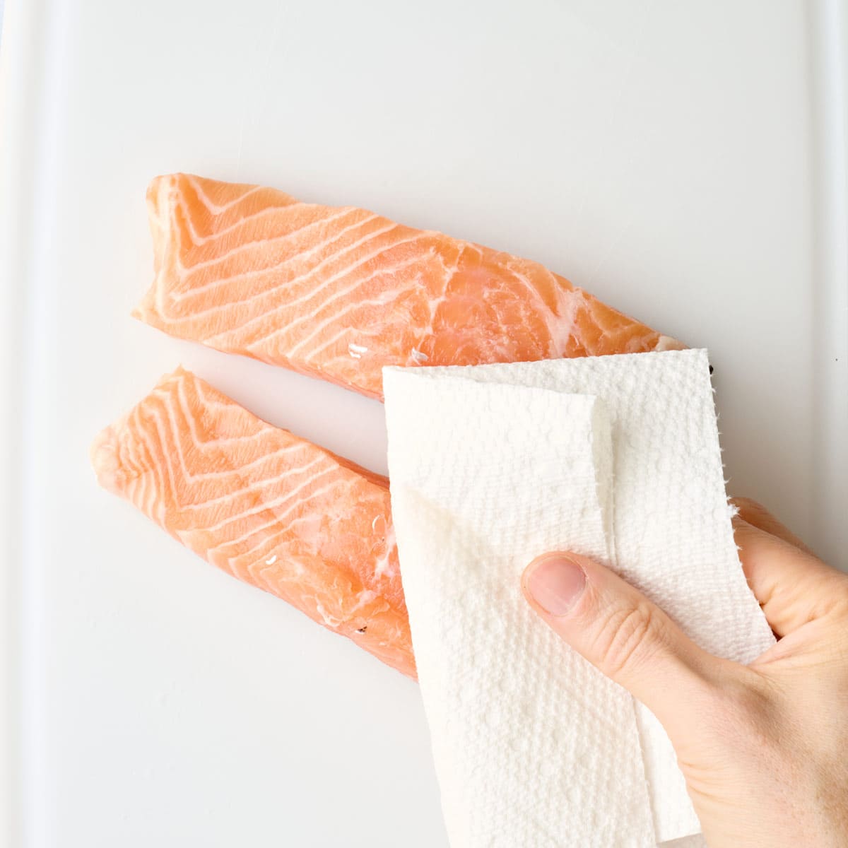 Salmon fillets on a white board being pat down with paper towels.