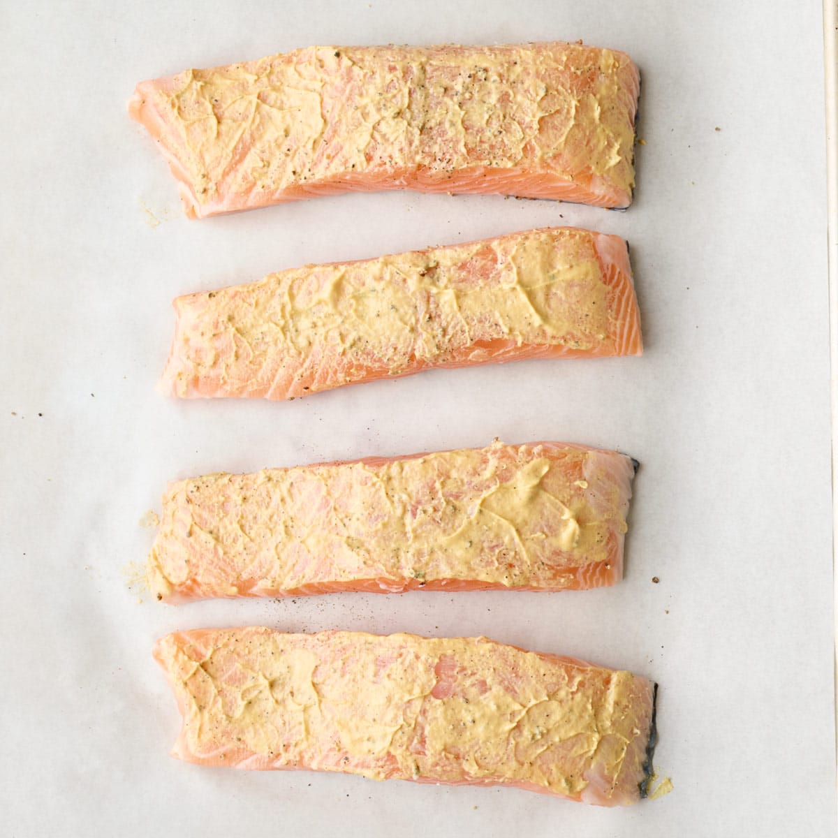 Salmon on a parchment lined sheet pan after seasoned with salt and pepper with Dijon mustard spread on top.